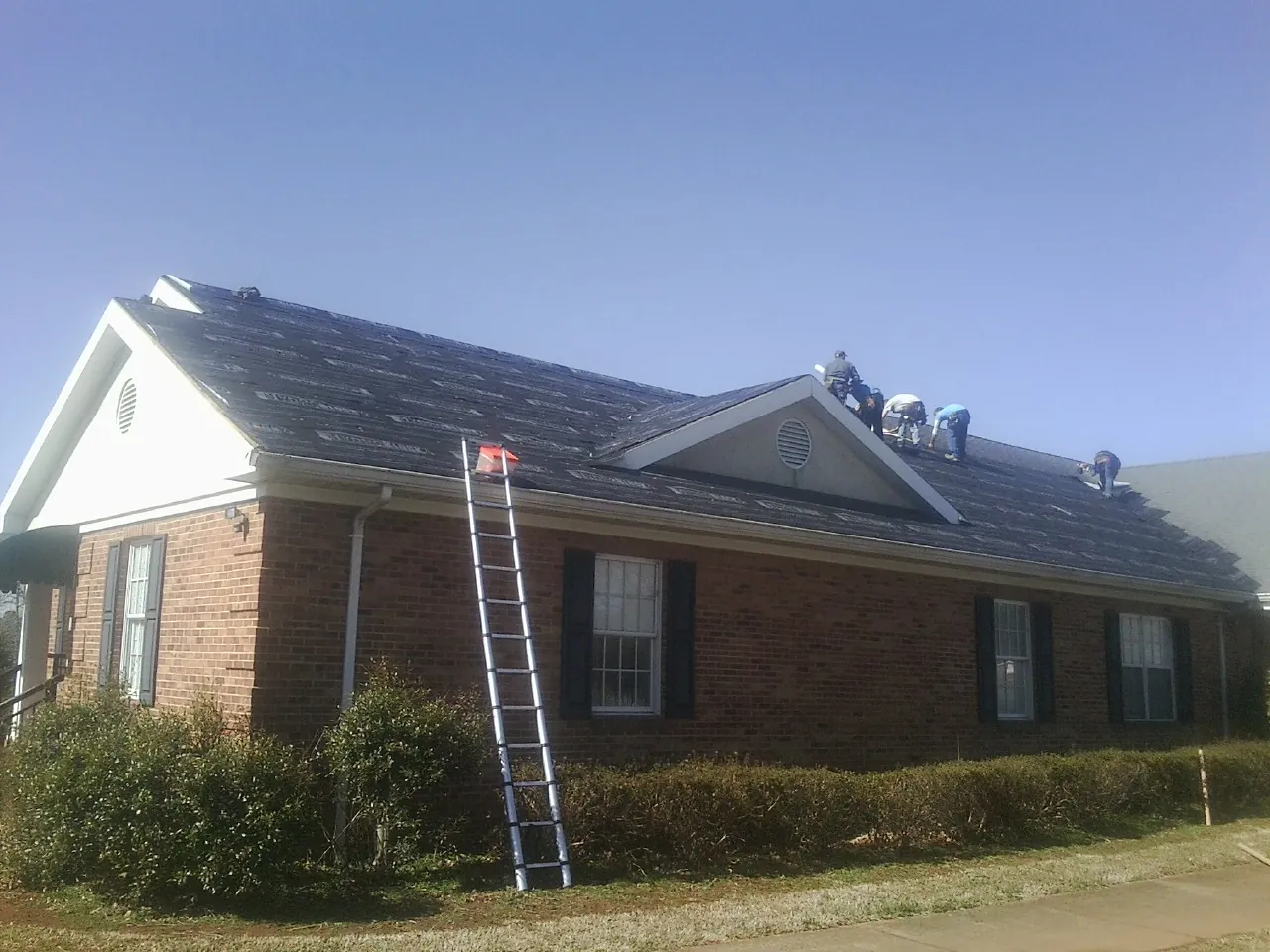 Crew tearing shingles off a brick ranch roof with a ladder set — Metal Roof Repair work by our crew in Sargeant