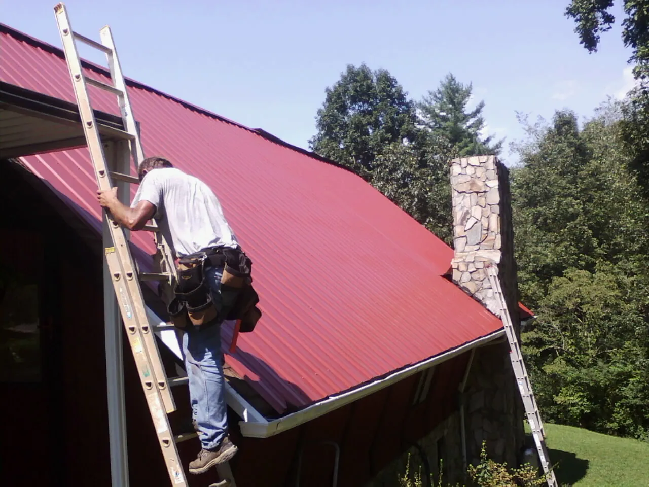 Roofer on a ladder beside a red metal roof and stone chimney — Soffit & Fascia Repair by Roofing Brothers in Sargeant